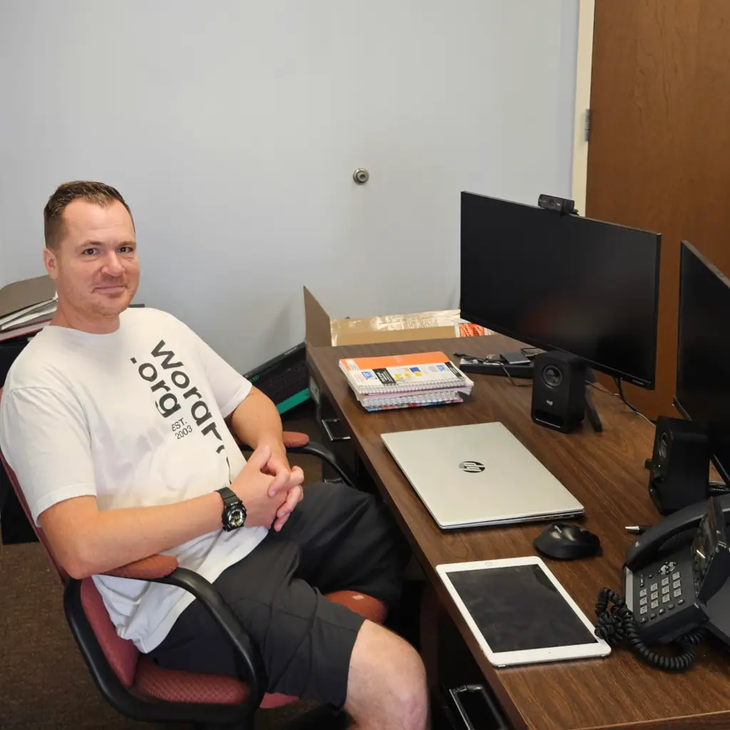Jesse Lange seated at office desk with monitor, laptop, tablet, and phone at Cherry Hill location.