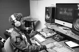 Black and white photo of student Hip Hop producer wearing headphones at music workstation with monitors & mixing equipment.