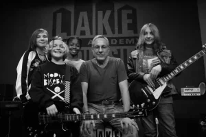 Black and white photo of Lakehouse Music Academy instructor with four young musicians holding guitars on stage at APMF academy program.