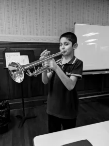 Black and white photo of a young boy playing trumpet in classroom with sheet music and whiteboard visible in background.