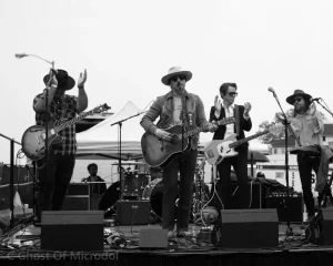 Black and white photograph of five musicians performing on outdoor stage with guitars, drums, and microphones.
