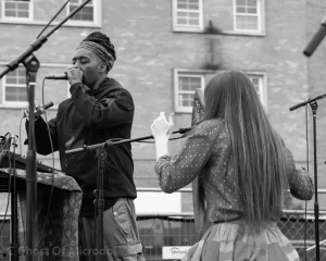Black and white photograph of Kuf Knotz performing at microphone with Christine Elise in an outdoor concert setting.