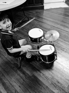 Young child smiling while playing a drum kit on wooden flooring in black and white photo.