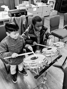 Black and white photo of two young boys holding drumsticks and practicing on a drum kit in a classroom setting.