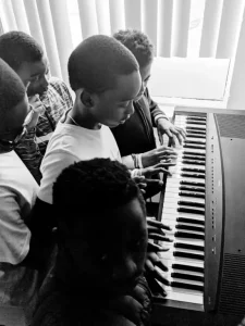 Multiple young children gathered around a digital keyboard, learning to play piano together in black and white photo.