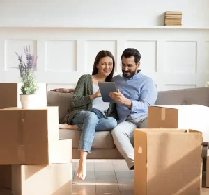 Smiling couple sitting on couch reviewing digital tablet surrounded by cardboard moving boxes in bright living room.